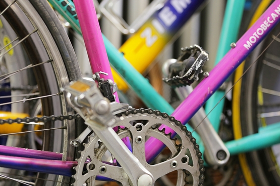 Eine Detailaufnahme der Pedale eines Violetten Rennrades. Im Hintergrund sind weitere bunte Fahrradrahmen zu sehen.