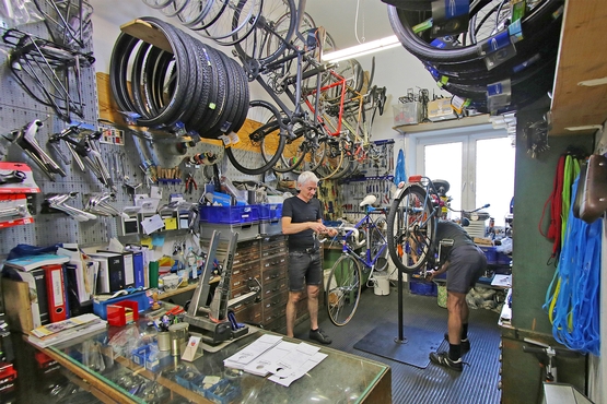 Ein Blick in die Werkstatt von klingeling! Im Hintergrund hängen Fahrräder, sowie Fahrradteile wie Reifen und Rahmen. Untern im Bild sind Mitarbeiter bei der Arbeit zu sehen.