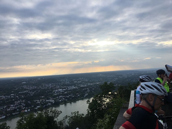 Mehrere Fahrradfahrer stehen an einem Geländer, im Hintergrund sieht man den Rhein.