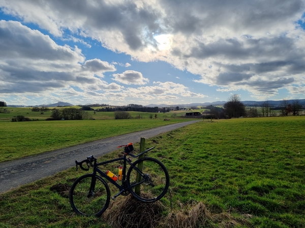 Eine Aussicht auf ein Fahrrad, das an einem Holzpfahl anlehnt, dahinter weite Felder und Gebirge im Hintergrund.